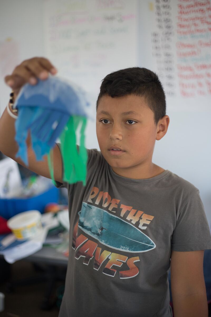 In the image, a young boy is holding up a homemade jellyfish. The jellyfish is made of blue and green materials. The boy is looking at the jellyfish with a thoughtful expression. He is wearing a gray t-shirt with a surfing graphic. The background is a classroom setting.